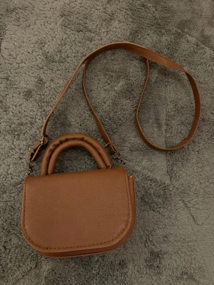 Brun minihandväska med axelrem - Snygg liten handväska i brunt konstläder med rundade former och vadderade handtag. Väskan har en lång justerbar axelrem och stängs med lock. Perfekt för dig som gillar minimalistisk och trendig stil.