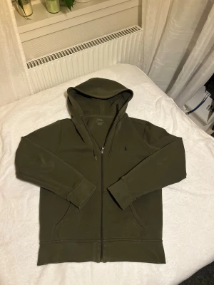 Olivgrön hoodie från Polo Ralph Lauren - Snygg olivgrön hoodie med dragkedja från Polo Ralph Lauren. Klassisk modell med huva och dragsnören, två fickor framtill och diskret logga broderad på bröstet. Tillverkad i mjuk bomullsblandning som känns skön mot huden. Perfekt för en avslappnad och stilren look.