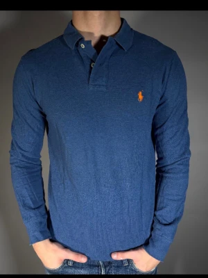 Blå långärmad skjorta från Ralph Lauren - Snygg blå långärmad skjorta från Ralph Lauren med klassisk krage och orange broderad logga på bröstet. Skjortan har knappar vid halsen och är gjord i mjuk bomull som känns skön mot huden. Perfekt för en clean och stilren look.