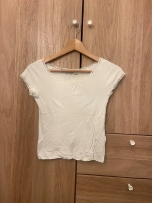 Vit topp med spetsdetaljer - Söt vit topp med korta ärmar och rund halsringning. Spetsdetaljer vid hals och ärmslut ger en romantisk touch. Figurnära passform och mjukt material, perfekt till jeans eller kjol.