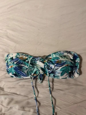 Blå blommig bikiniöverdel med knyt - Snygg bikiniöverdel från Hollister med tropiskt blommigt mönster i blått, grönt och orange. Bandeau-modell med knyt framtill och avtagbara axelband. Har aldrig använt den!