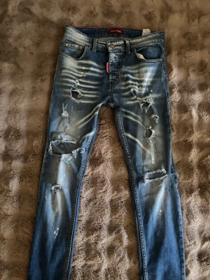 Blå slitna jeans från Dsquared2 - Säljer ett par blå jeans från Dsquared2 med coola slitningar och hål på benen. Jeansen har slim fit, klassisk femficksmodell och detaljerade slitningar för en trendig look. Materialet är denim i bomull. Perfekta för dig som gillar streetstyle. Obs A kopia