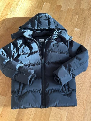 Svart pufferjacka från Urban Classics - Snygg svart pufferjacka från Urban Classics med huva och dragkedja framtill. Jackan har en quiltad design, hög krage och två sidofickor. Perfekt för kalla dagar och ger en riktigt trendig look. Materialet är slitstarkt och håller värmen bra. Huvan är avtagbar. Jackan är så gott som ny i princip aldrig använd förutom testat!🙌 Hör av er vid frågor eller andra funderingar✅ Storlek 164 som motsvarar ca xs😆