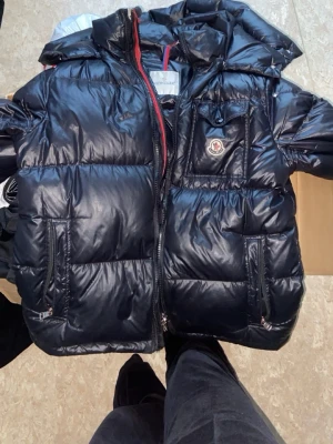 Svart dunjacka från Moncler - Snygg svart dunjacka från Moncler med glansig finish, huva och dragkedja framtill. Jackan har två framfickor med dragkedja och en bröstficka med Moncler-logga. Insidan har ett unikt serietryck. Storlek M. Perfekt för kalla vinterdagar.  Vid snabb affär kan priset diskuteras