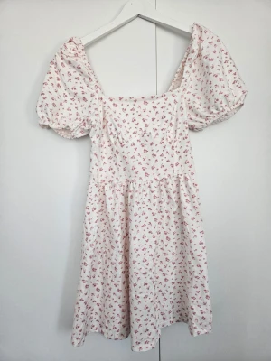 Vit blommig kortklänning med puffärm - Söt vit klänning med små rosa blommor och puffiga kortärmar. Klänningen har en fyrkantig urringning och är gjord i mjuk bomull. Den har en avslappnad passform och romantisk känsla.