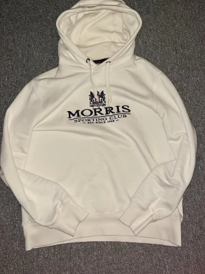 Beige Morris hoodie - Snygg beige hoodie från Morris Sporting Club med broderad logga och text på bröstet. Klassisk huva med dragsko och mjukt material på insidan. Perfekt för en avslappnad och stilig look. Tröjan passar dig från 165~173cm, har du några frågor är det bara att skriva