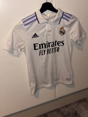 Real Madrid matchtröja Adidas vit - Vit Real Madrid matchtröja från Adidas med lila detaljer på axlarna och svart text. Tröjan har klubbmärke, Adidas-logga och 'Emirates Fly Better' tryckt på bröstet. Klassisk pikékrage och korta ärmar. Perfekt för fotbollsträning eller att bära på matchdag. Den har 2 fläckar som inte går bort o tvätten men ni kanske kan lösa det! 