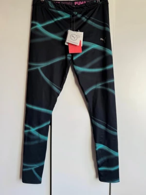 Nya träningsleggings  kompression. Puma. L. NWT. - Snygga svarta träningsleggings från Puma med turkosa vågiga linjer och diskret Puma-logga på benet. Tillverkade i ett stretchigt funktionsmaterial med avancerad kompression (CELL Level 3) för extra stöd vid träning. Perfekta för gymmet eller löprundan. Oanvända. Alla lappar kvar. Storlek L. Finns inget att anmärka på. NWT.