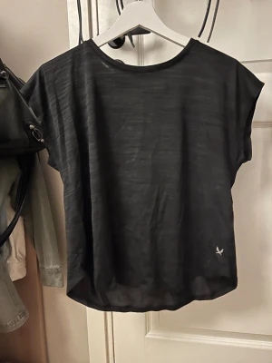 Svart sport-topp från SOC - En svart sport-topp från SOC med rund hals och korta ärmar. Tillverkad i ett tunt, tekniskt material som andas, perfekt för träning eller gym. Diskret logga nertill. Enkel och stilren design som passar till det mesta inom sport.