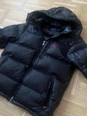 Svart dunjacka från Polo Ralph Lauren - Snygg svart dunjacka från Polo Ralph Lauren med huva och dragkedja framtill. Jackan har två stora fickor med dragkedja och den klassiska röda loggan broderad på bröstet. Puffig modell som håller dig varm under kalla dagar.