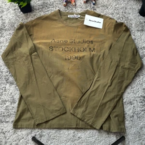 Olivgrön långärmad topp Acne Studios - Olivgrön långärmad topp från Acne Studios med tryckt text 'STOCKHOLM 1996' framtill. Tillverkad i mjuk bomull med avslappnad passform och rund halsringning. Perfekt för en chill och stilren look.