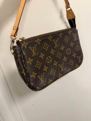 Lv väska - Snygg brun handväska från Louis Vuitton med klassiskt monogrammönster och guldfärgade detaljer. Väskan har en avlång form, dragkedja upptill och en beige läderrem. Perfekt för dig som gillar lyxiga accessoarer och tidlös design.