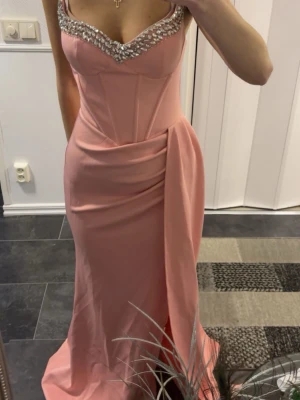 Bal klänning  - Elegant långklänning i rosa med figurnära passform och vackra pärldetaljer längs urringningen. Klänningen har en draperad detalj vid höften och markerad midja för en snygg siluett. Perfekt för speciella tillfällen. Aldrig använd 