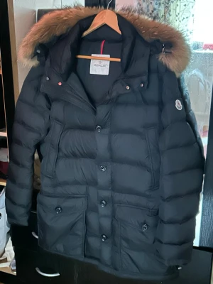 Svart Moncler dunjacka med pälskrage - Snygg svart dunjacka från Moncler med stor huva och pälskant. Jackan har knappar framtill, två stora fickor med lock och klassisk Moncler-logga på ärmen. Perfekt för kalla vinterdagar och riktigt varm tack vare dunfyllningen. Passar M-L