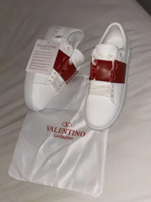 Valentino Garavani Open - sneakers, vinröd/vit - Helt nya, fräscha Valentino Garavani Open sneakers i vitt skinn med en bred vinröd läderdetalj över mitten. Klassisk rund tå, platt sula och coola nitar baktill på sulan. Skorna har vita snören och diskret logga på plösen. Perfekta för dig som gillar lyxig streetstyle.