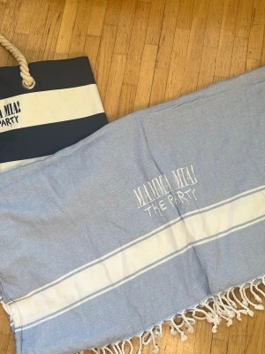 Blå strandhandduk Mamma Mia! The Party - Säljer en ljusblå strandhandduk med vita ränder och fransar i kanterna. Handduken har broderad text 'Mamma Mia! The Party' och är tillverkad i mjukt tyg, perfekt för stranden. Medföljer en matchande blåvit strandväska med rep-handtag.