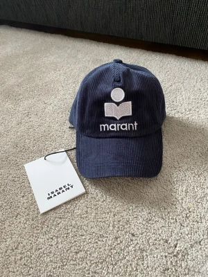Isabel Marant keps i Manchester/Corduroy - Snygg marinblå keps från Isabel Marant i bredspårig manchester. Kepsen har böjd skärm och vit broderad logga framtill. Justerbar baktill för perfekt passform. Perfekt accessoar för en cool och avslappnad stil.