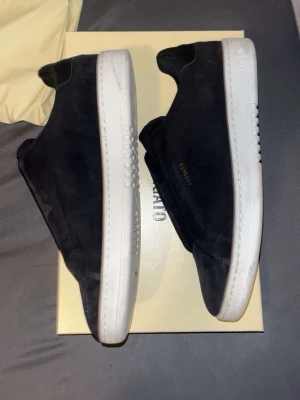 Svarta Axel Arigato sneakers - Stilrena svarta slip-on sneakers från Axel Arigato med vit sula och minimalistisk design. Skorna har diskret guldfärgad logga på sidan och är tillverkade i mocka. Perfekta för dig som gillar clean och modern stil. Nästan helt ny och bara använd 2-3 gånger. Original pris är 3000 kronor