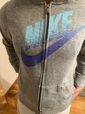 Grå zip hoodie från Nike - Snygg grå hoodie från Nike med dragkedja och stor logga i blått och turkost framtill. Hoodien har huva och två fickor på magen. Tillverkad i mjukt sweatshirtmaterial som är skönt att ha på sig. Perfekt för en avslappnad och sportig stil.