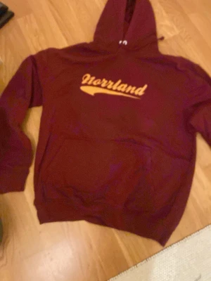 Vinröd hoodie med Norrland-tryck - Säljer en vinröd hoodie med gul text där det står 'Norrland' på bröstet. Hoodien har klassisk huva och magficka framtill. Perfekt för dig som vill ha en chill och snygg stil med lite norrländsk vibe. Materialet känns mjukt och skönt.