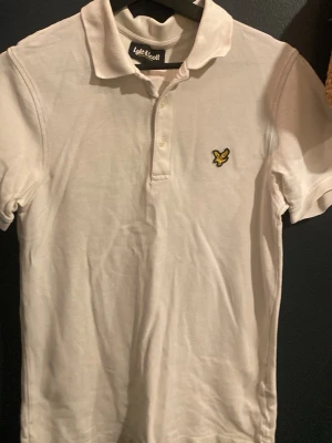 Vit pikétopp från Lyle & Scott - Säljer en klassisk vit pikétopp från Lyle & Scott med krage och knappar framtill. T-shirten har korta ärmar och den ikoniska gula logon på bröstet. Perfekt för dig som gillar stilrena och sportiga plagg.