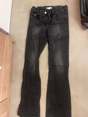 Svarta låg midjade bootcut jeans - Snygga svarta jeans med bootcut passform. Klassisk femficksmodell med två bakfickor med lock och silverfärgade knappar. Jeansen har dragkedja och knapp i midjan. Perfekta för en avslappnad och trendig look. Går även att tajta åt i midjan. 