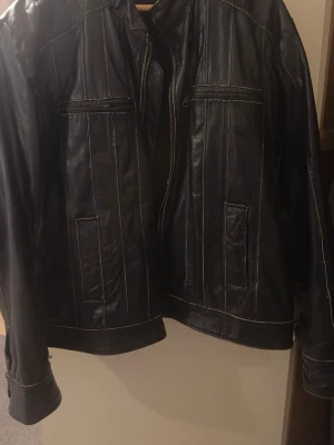 Svart skinnjacka från Woodland XL - Snygg svart skinnjacka från Woodland i storlek XL. Jackan har dragkedja framtill, två bröstfickor med dragkedja och två sidofickor. Klassisk rak passform och markerade sömmar ger en cool look. Perfekt för dig som vill ha en tidlös och stilren jacka.