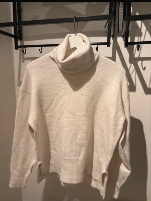 Beige polotröja med slitsar - Mysig beige stickad polotröja med breda ribbade muddar och höga slitsar i sidorna. Tröjan har en lös passform och hög polokrage som ger en avslappnad look. Perfekt för lager på lager och höstmys.