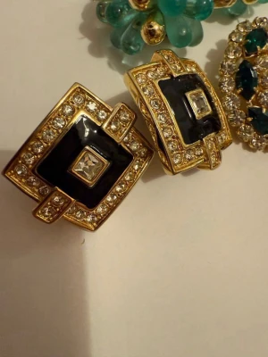 Svart och guldiga statementörhängen - Säljer ett par fyrkantiga statementörhängen i svart och guld med gnistrande stenar runt kanterna och i mitten. Örhängena har en lyxig vintagekänsla och är perfekta för dig som gillar att sticka ut. Metall och emalj i kombination med klara stenar ger en exklusiv look.