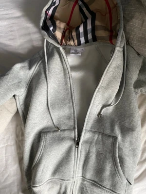 Grå zip hoodie från Burberry - Snygg grå zip hoodie från Burberry med klassiskt rutigt foder i huvan och dragsko. Hoodien har två stora fickor framtill och ribbade muddar.  Säljer pågrund av att den är för liten för mig så om du är S skulle jag tro den passar bättre på dig..