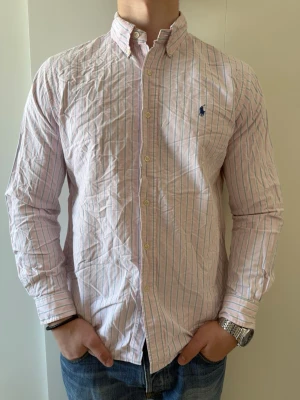 Rosa randig skjorta från Ralph Lauren - Klassisk rosa skjorta från Ralph Lauren med tunna blå och vita ränder. Skjortan är i storlek L och mycket bra skick utan defekter 