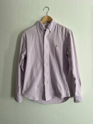 Ljuslila skjorta från Polo Ralph Lauren - Säljer en ljuslila skjorta från Polo Ralph Lauren i stretchig oxford-bomull. Skjortan har klassisk krage, knappstängning framtill, lång ärm och den ikoniska broderade loggan på bröstet. Slim fit-modell som sitter snyggt och stilrent.