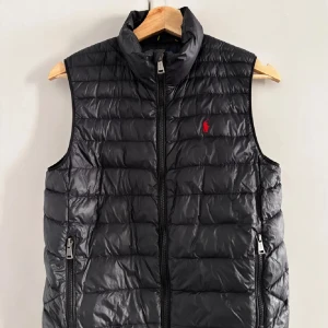 Svart dunväst från Ralph Lauren - Snygg svart dunväst från Ralph Lauren med dragkedja och två fickor. Den har en diskret röd logga på bröstet och är perfekt för kyligare dagar. Passar 160-164