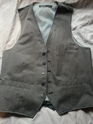 Grå kostymväst från Skill - Snygg grå kostymväst från Skill i storlek M. Västen har tunna vita ränder, klassisk v-ringning och fem knappar framtill. Fodret är också randigt och västen har tre fickor fram. Perfekt för en stilren och dressad look.
