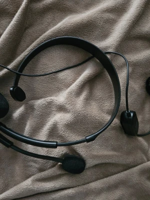Svart trådbundet headset med mikrofon - Säljer ett svart headset med tunn bygel och inbyggd mikrofon på armen. Headsetet har öronkuddar i skum och ansluts med sladd. Perfekt för gaming, samtal eller distansstudier. Enkel och lätt design i plast.