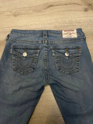 Blå True Religion jeans med fickdetaljer - Säljer ett par blå jeans från True Religion med ikoniska kontrastsömmar och stora ficklock bak. Jeansen har silverfärgade knappar och en patch med logga i midjan. Klassisk rak passform och slitna detaljer för en avslappnad look.