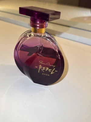 Avon Far Away Rebel parfym - Säljer en snygg parfym från Avon, Far Away Rebel. Flaskan är lila med en unik, rundad form och har ett fyrkantigt lock i mörklila plast. Guldig detalj vid halsen ger en lyxig känsla. Perfekt för dig som gillar dofter med attityd. 75% kvar. 
