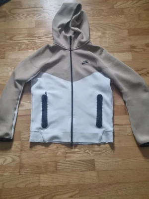 Nike beige och vit hoodie - Säljer en snygg hoodie från Nike i beige och vitt med svarta detaljer. Hoodien har dragkedja framtill, två svarta fickor med dragkedja och en klassisk Nike-logga på bröstet. Tillverkad i mjukt material och har en stor huva för extra komfort.