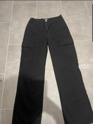 Svarta cargobyxor med fickor - Säljer ett par svarta cargobyxor med raka ben och stora fickor på sidorna. Byxorna har knapp- och dragkedjestängning samt extra fickor baktill. Perfekta för en avslappnad och trendig look. Materialet känns slitstarkt och passar till streetwear.