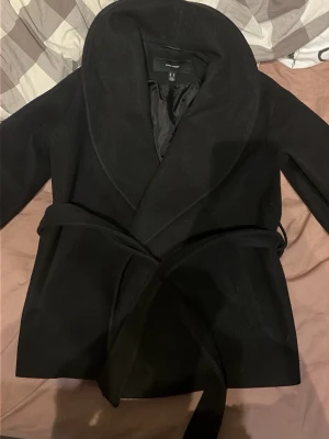 Svart kappa från H&M med skärp - Stilren svart kappa från VERO MODA med bred krage och knytband i midjan. Kappan har en klassisk, rak passform och är fodrad på insidan för extra komfort. Perfekt för kyliga dagar och enkel att matcha med olika outfits.