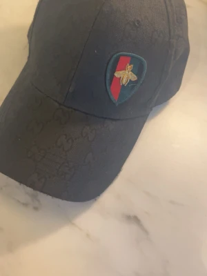 Svart keps från Gucci med bi-detalj - Snygg svart keps från Gucci med diskret GG-mönster i tyget och en patch framtill med röd och grön rand samt en guldfärgad bi. Justerbart spänne bak i metall med Gucci-logga. Perfekt accessoar för dig som gillar exklusiv streetstyle.