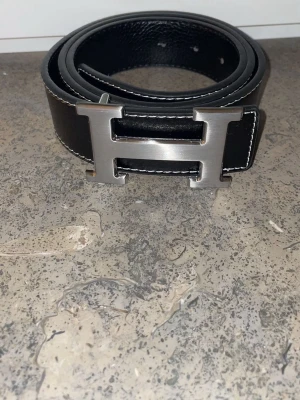 Svart bälte med H-spänne - Snyggt svart bälte med det ikoniska H-spännet i borstat silver. Bältet har vita kontrastsömmar längs kanterna och är gjort i slätt skinn. Perfekt accessoar för att lyfta vilken outfit som helst. Tveka inte att höra av er vid frågor eller funderingar 🤗