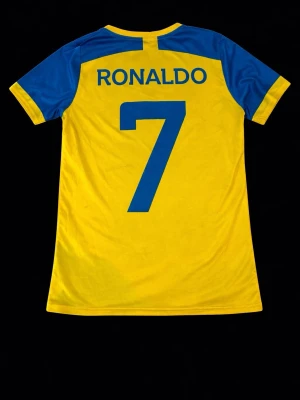 Al Nassr Ronaldo matchtröja gul/blå - Snygg gul matchtröja från Al Nassr med blå detaljer och tryck. På ryggen står det 'Ronaldo' och nummer 7 i blått. Framsidan har klubbmärke och sponsorlogga i blått. Perfekt för dig som älskar fotboll och vill stötta ditt favoritlag.