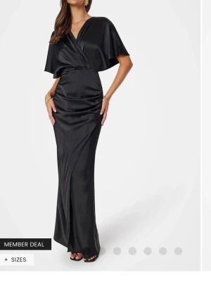 Svart långklänning i satin - Elegant svart långklänning i glansigt satinmaterial med omlott upptill och v-ringad hals. Klänningen har korta, vida ärmar och markerad midja som ger en snygg siluett. Perfekt för dig som vill sticka ut på nästa event.