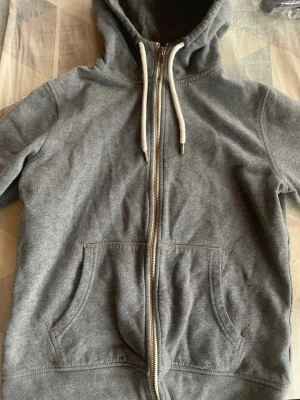 Grå zip hoodie från The Essentials - Mysig grå hoodie från The Essentials med dragkedja framtill, vita dragsnören och två stora fickor. Tillverkad i ekologisk bomull och har en klassisk huva. Perfekt för chill dagar eller när du vill ha en enkel och stilren look. 🩶