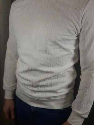 Vit stickad tröja med mönster - Säljer en vit stickad tröja med diskret, upprepat mönster i samma färg. Tröjan har rund halsringning, långa ärmar och ribbade muddar vid ärmslut och nederkant. Perfekt för dig som gillar stilrena detaljer och en modern look.