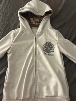 Vit hoodie med leopardfoder Challenge - Vit hoodie med dragkedja från Challenge. Huvan är fodrad med leopardmönstrat, mjukt tyg. Tryck med Challenge-logga och lejon på bröstet. Hoodien har ribbade muddar och är tillverkad i ett mjukt material som känns skönt mot huden.