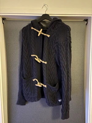 Mörkblå stickad kofta med huva Ralph Lauren - Mörkblå grovstickad kofta från Denim & Supply Ralph Lauren med huva och snygga snörningsdetaljer i beige. Koftan har två fickor framtill och coola träknappar. Storlek S men passar även M
