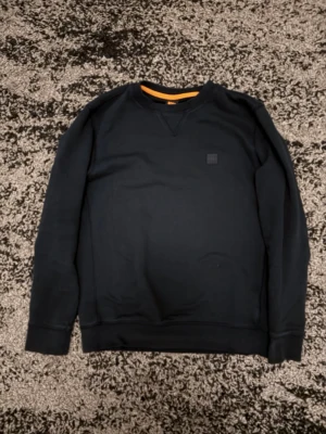 Svart sweatshirt från Hugo Boss - Klassisk svart sweatshirt från Hugo Boss med rund halsringning och diskret logopatch på bröstet. Tröjan har ribbade muddar vid ärmslut och nederkant samt en orange detalj på insidan av kragen. Perfekt för en clean och stilren look.