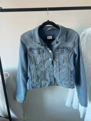 Ljusblå klassisk jeansjacka - Säljer en ljusblå jeansjacka med klassisk krage, silverfärgade knappar och två bröstfickor med lock. Jackan har en rak passform och snygga sömdetaljer framtill. Jackan är i barnstorlek men passar som en xs/s.